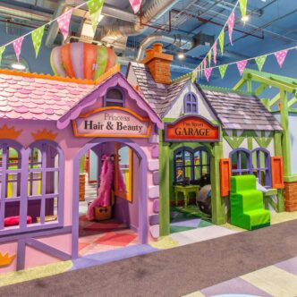 Purple soft play house with a sign that reads 'Princess Hair and Beauty' next to a green soft play house with a 'Garage' sign. Green and pink polka dot bunting hangs above.