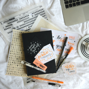 A black journal surrounded by orange pens. Handwritten cursive notes read: "Grow positive thoughts" and "A better me is coming".