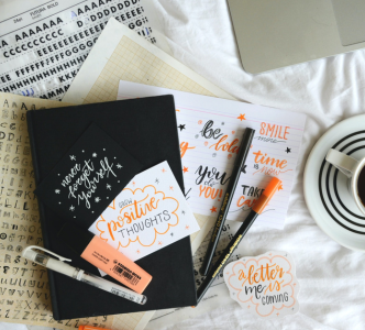 A black journal surrounded by orange pens. Handwritten cursive notes read: "Grow positive thoughts" and "A better me is coming".