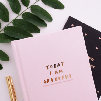 A pink journal with "Today I am grateful" embossed in gold on the front sitting on top of a black notebook, next to a green leaf